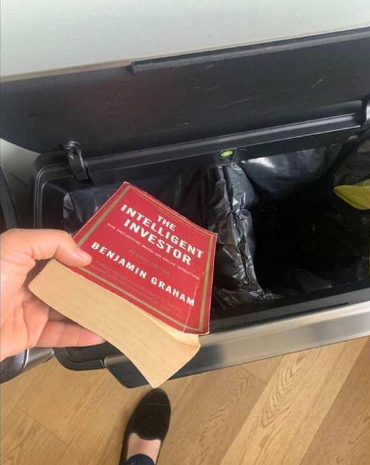 Une main tenant le livre The Intelligent Investor de Benjamin Graham au-dessus d'une poubelle ouverte